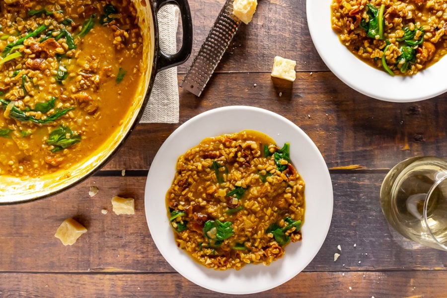 a white plate with farro risotto on top of a wooden table surrounded by chunks of scattered parmesan cheese, a second plate of risotto and a large pan of risotto