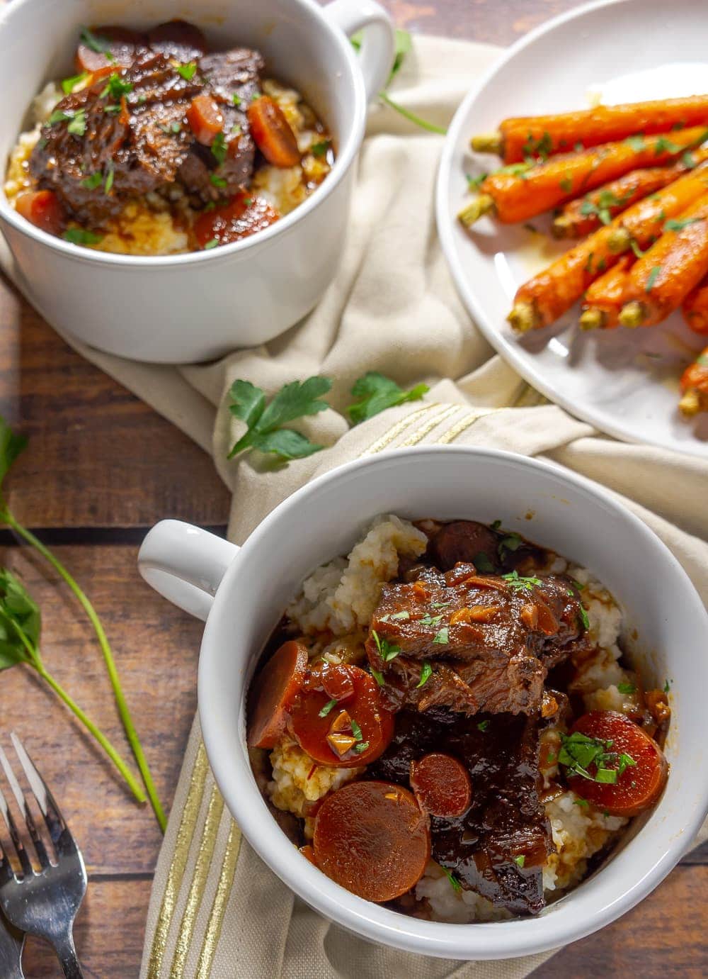 two white bowls of braised beef on a wooden table next to a plate of orange carrots