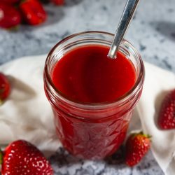 A jar of strawberry syrup surrounded by fresh strawberries