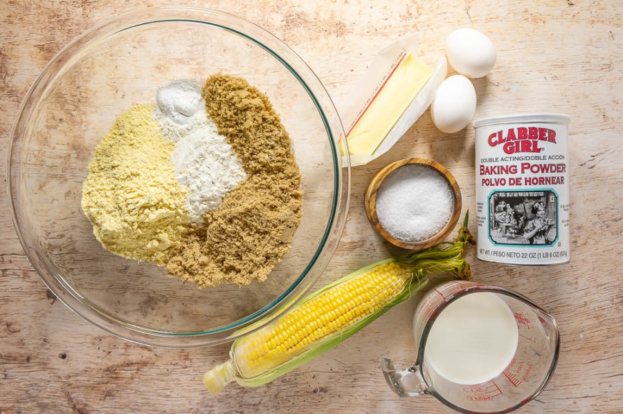 bowl of flour, cornmeal, and brown sugar with butter salt, eggs, baking powder, milk, and corn beside it