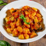 gnocchi in tomato sauce in a while bowl on a wooden table surrounded by basil and fresh tomatoes
