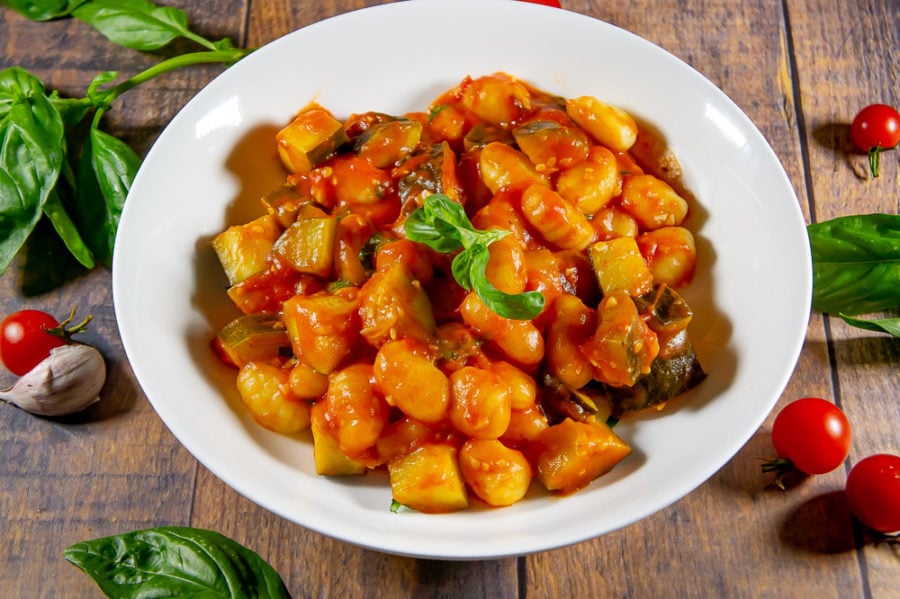 gnocchi in tomato sauce in a while bowl on a wooden table surrounded by basil and fresh tomatoes