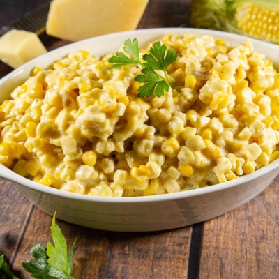an oval bowl of creamy corn on a wooden table surrounded by parsley, parmesean cheese and a corn cob