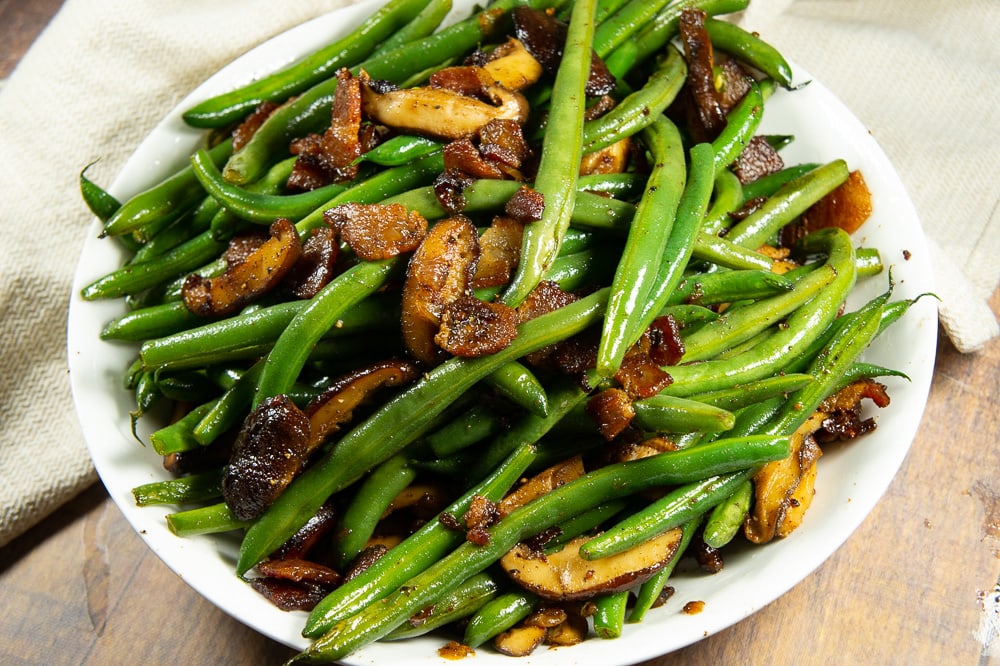 a circular white bowl full of green beans with bacon on a wooden table next to a white towel