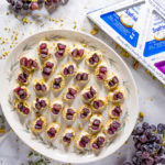 a platter of bite sized crackers topped with goat cheese, pistachios and grapes surrounded by thyme, grape clusters and goat cheese packaging