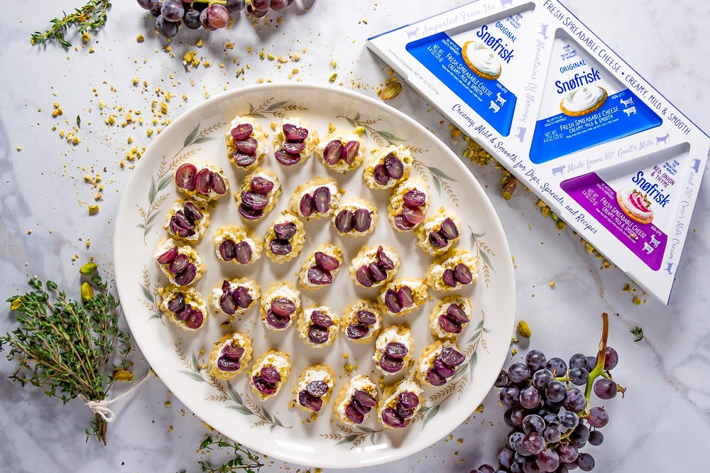 a platter of bite sized crackers topped with goat cheese, pistachios and grapes surrounded by thyme, grape clusters and goat cheese packaging
