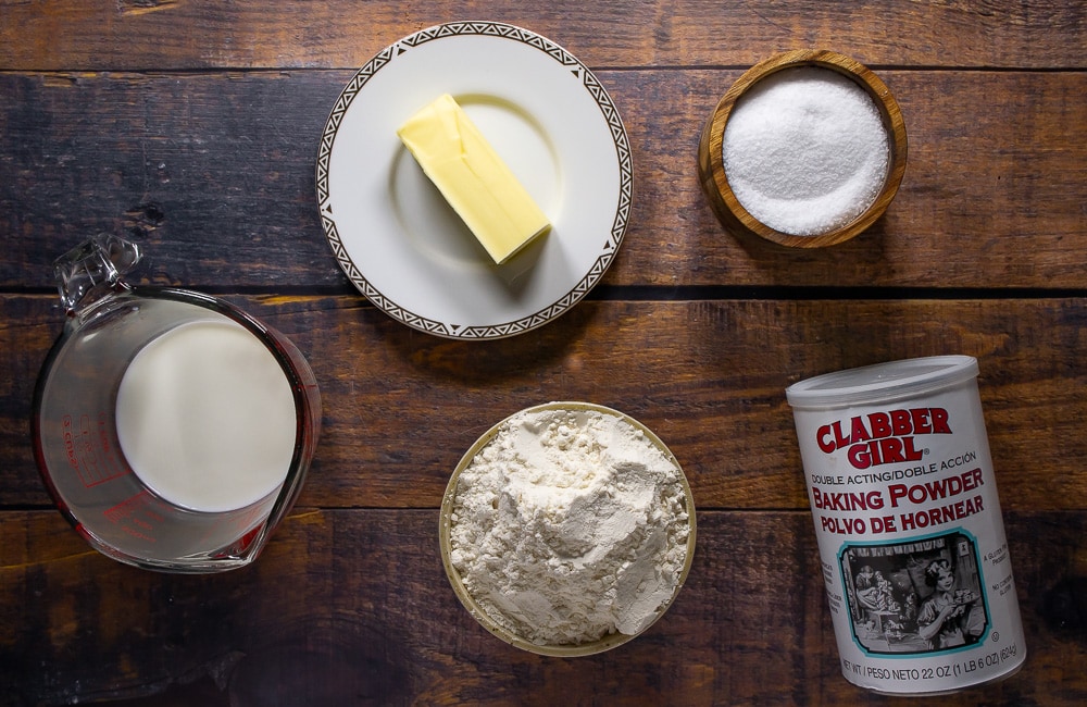 a bowl of flour, a stick of butter, a cup of milk, salt, and baking powder on a wooden table