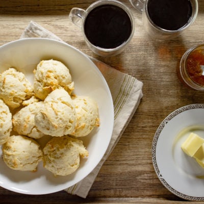 a bowl of drop biscuits on a wooden serving tray with two cups of coffee, jam, and butter