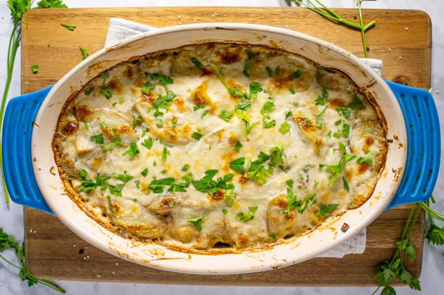 a blue casserole dish with golden brown scalloped potatoes topped with parsley on a cutting board on a white granite counter