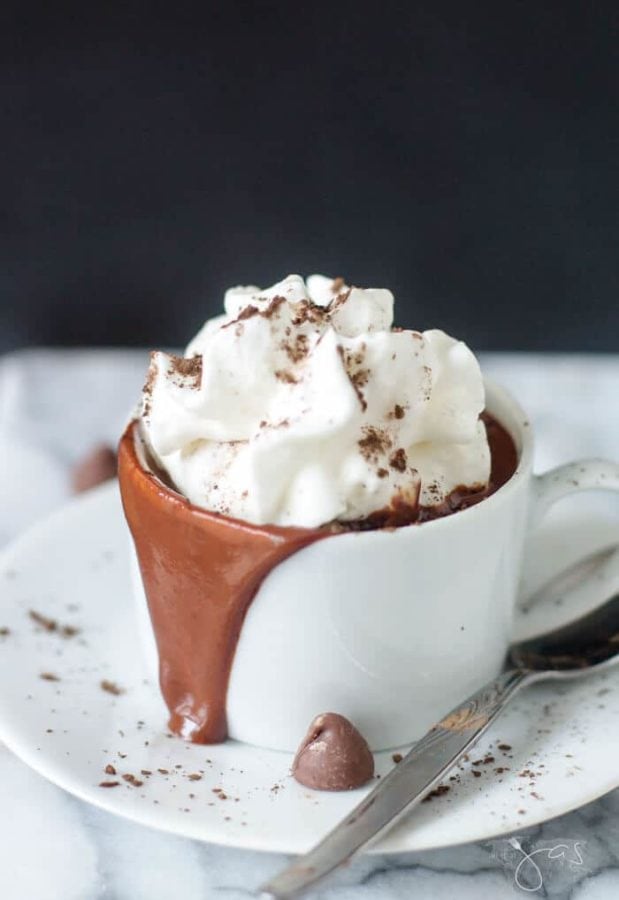a white cup with drinking chocolate inside and spilling over the rim onto a white saucer topped with whipped cream.