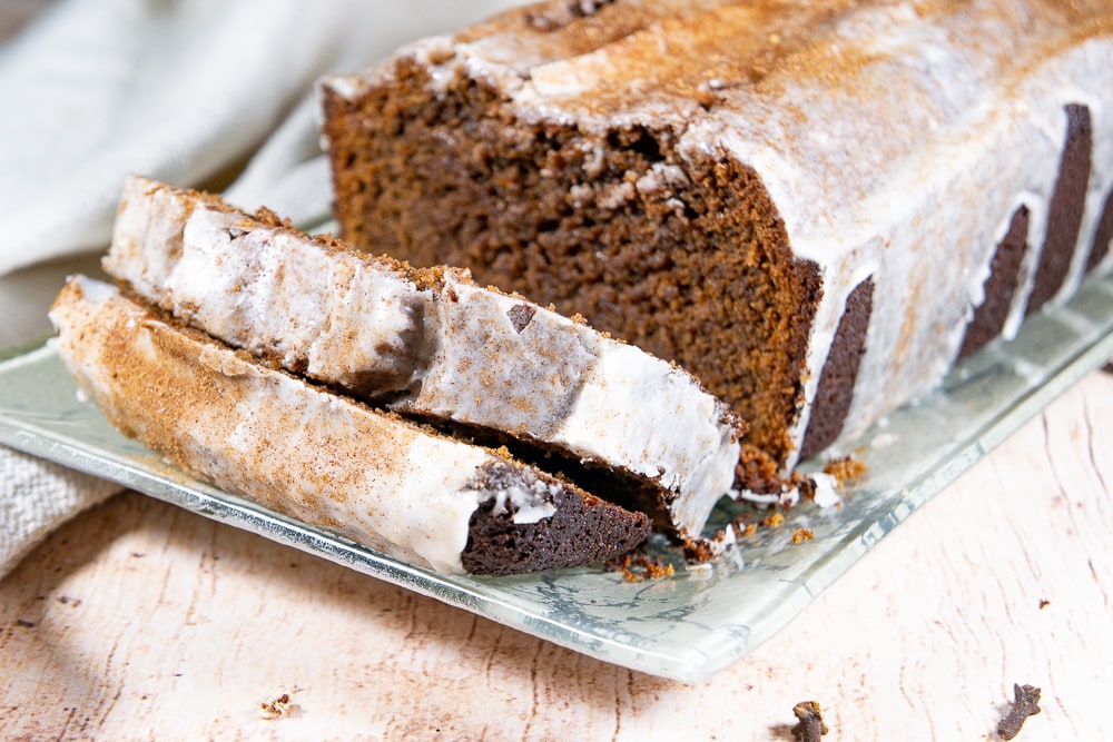 A loaf of ginger and chocolate quick bread with icing, on a green platter with two slices cut from the front and laying on the platter