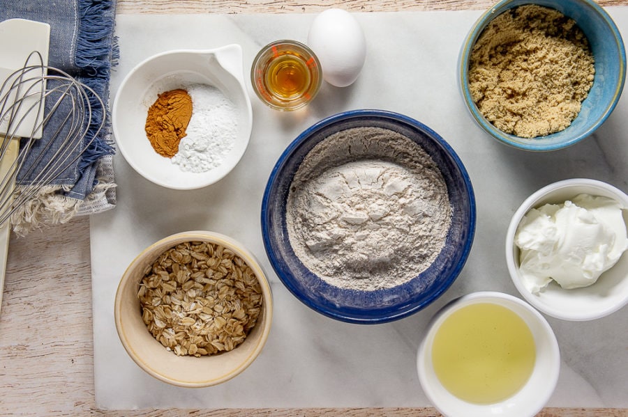 A white granite cutting board with a blue bowl of wheat flour, a blue bowl of brown sugar, a white bowl of yogurt, a white bowl of spices, a glass of vanilla, a brown bowl of oats, and a white bowl of oil sitting atop a wooden table with a blue fringed napkin next to it.