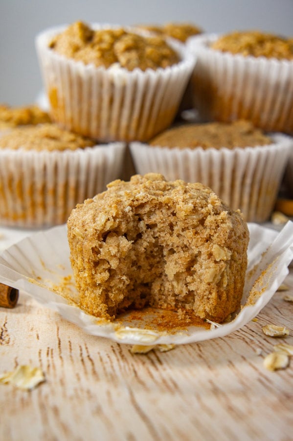 A stack of oatmeal muffins on a white cutting board behind a single muffin on a wooden table unwrapped and a bite taken out of it. 