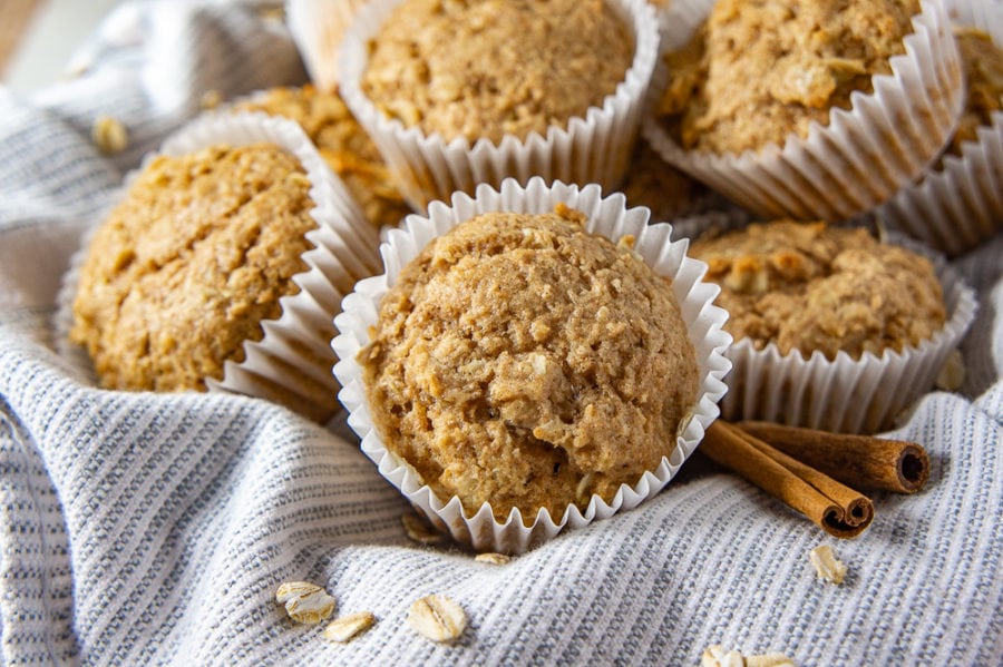 A pile of oatmeal cinnamon muffins on a blue and white towel with scattered oats and cinnamon sticks