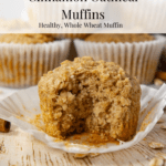A stack of oatmeal muffins on a white cutting board behind a single muffin on a wooden table unwrapped and a bite taken out of it.