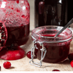 A small flip top jar of cranberry pomegranate jam with open pomegranates and cranberries around it in front of several larger jars of the jam