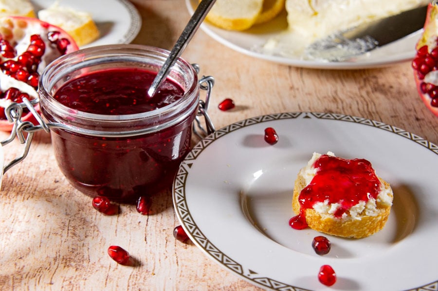 An open jar of cranberry jam with pomegranate beside a white plate with a brie crostini and jam drizzled on top of it.