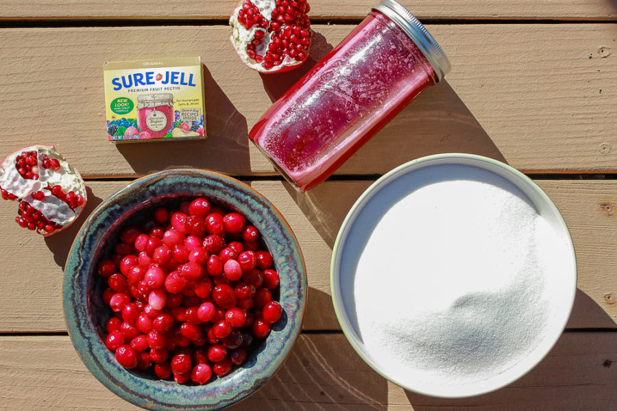 A bowl of cranberries, a bowl of sugar, and a jar of pomegranate juice with a box of pectin on a wood surface