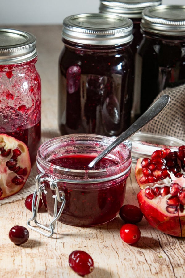 A small flip top jar of cranberry pomegranate jam with open pomegranates and cranberries around it in front of several larger jars of the jam