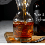 homemade vanilla extract being poured from a large amber mason jar into a decanter on a wooden cutting board beside a whole vanilla bean