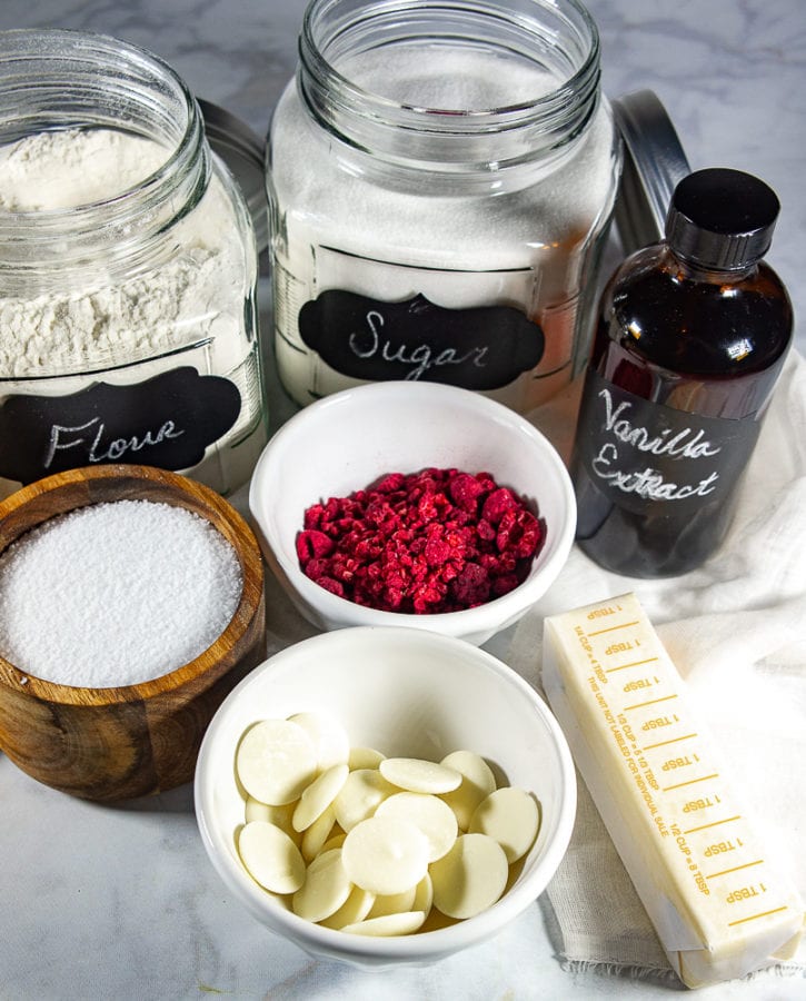 A white granite table with a glass jar of flour, a glass jar of sugar, a bottle of vanilla extract, a bowl of freeze dried raspberries, butter, white chocolate, and salt