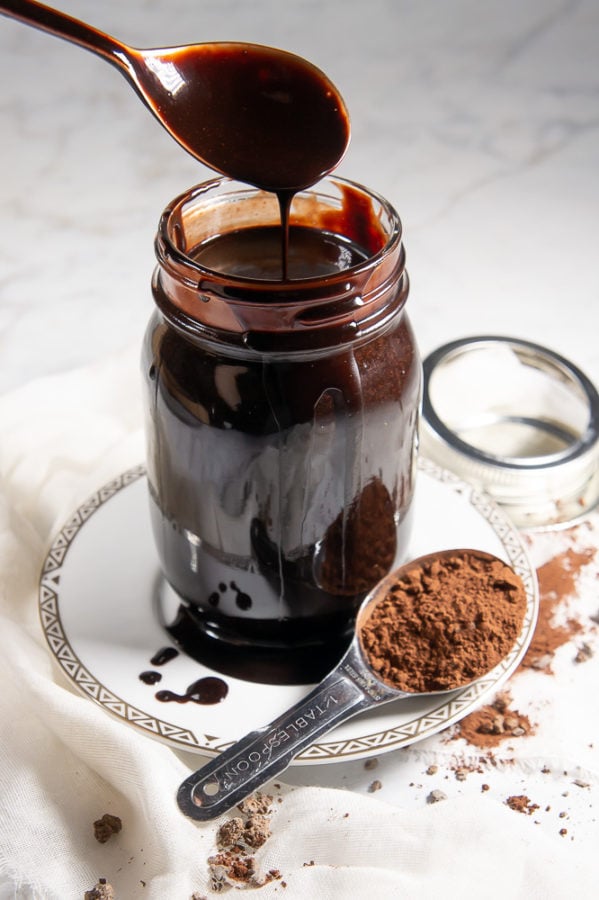 a white plate on a granite surface with a spoon of cocoa powder spilled around it. A jar of chocolate sauce on the plate with a spoon being lifted out of it.