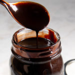 a spoon coated in chocolate syrup dripping into a jar of chocolate on a white granite surface