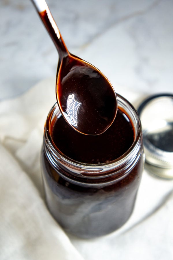 A glass jar with chocolate syrup and a spoon coated with chocolate syrup held above it surrounded by white cloth on a white granite surface