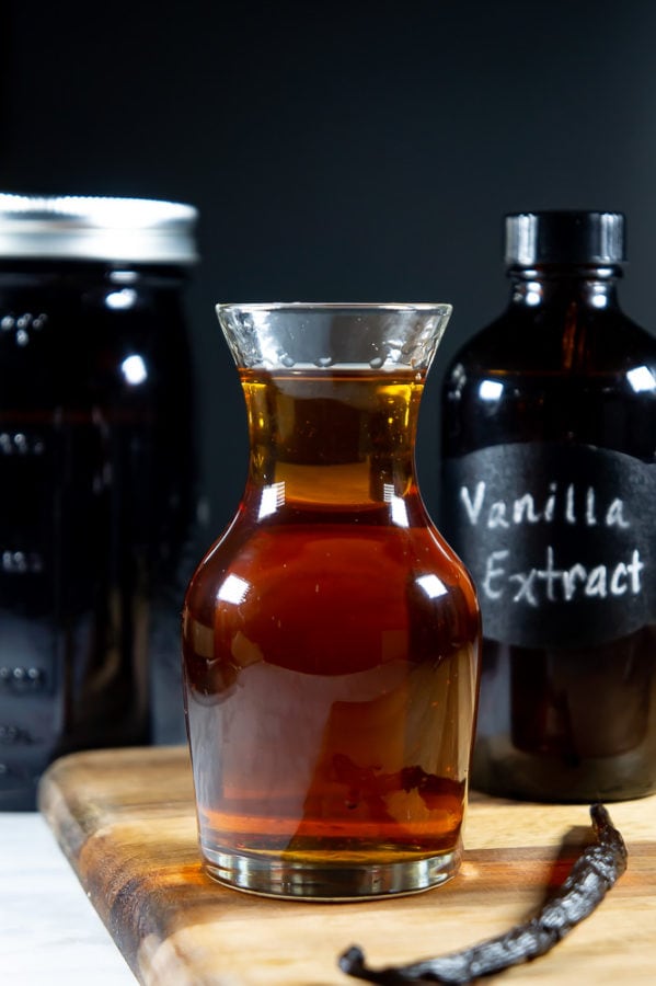 a decanter of vanilla extract on a wooden cutting board, a vanilla bean beside it, and a dark bottle of extract behind it.