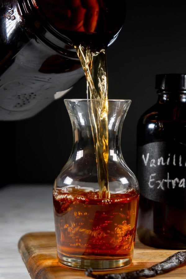 homemade vanilla extract being poured from a large amber mason jar into a decanter on a wooden cutting board beside a whole vanilla bean