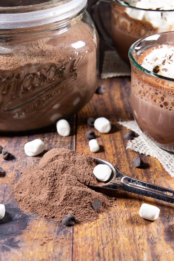 A large glass jar of hot cocoa mix with a spoonful scattered across the wooden table in front of it sitting beside two glasses of hot cocoa with whipped cream