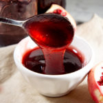 a white bowl with red pomegranate jelly being spooned in on a tan napkin in front of several jars of pomegranate jelly and a half pomegranate