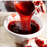 a white ramekin of pomegranate jelly with a spoon drizzling jelly into it with an open pomegranate beside it