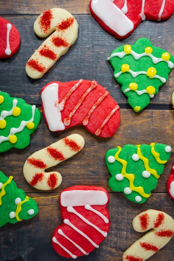 Three candy cane sugar cookies with red sprinkles, four green christmas tree sugar cookies, and four red stocking sugar cookies on a dark wooden table