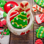 a santa towel beside a muffin cookie jar with sugar cookies and a cooling rack with cookies on a wooden table