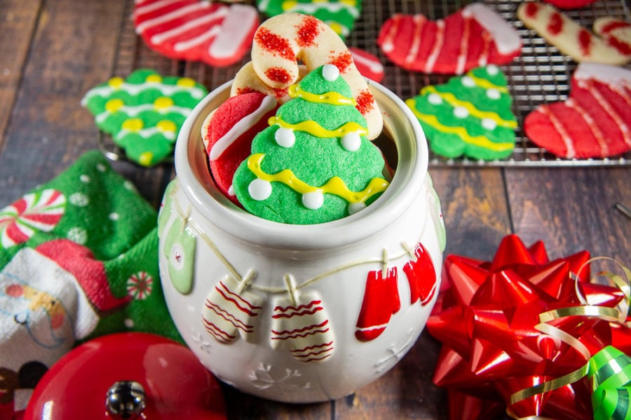 a white christmas cookie jar brimming with colored sugar cookies on a wooden table in front of a metal cooling rack with more cookies