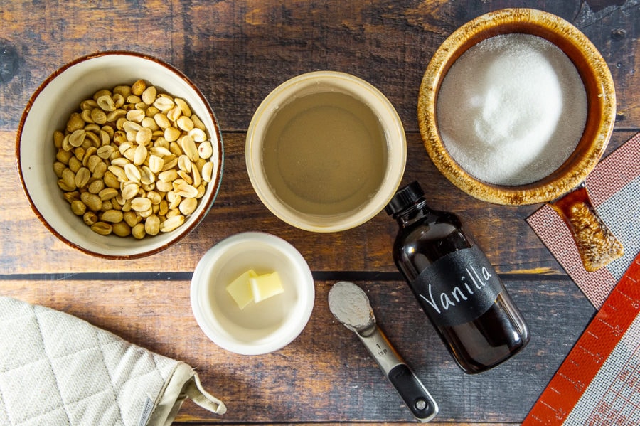 Ingredients for peanut brittle: a bowl of peanuts, a bowl of sugar, a bowl of karo syrup, a teaspoon of butter, and baking soda, and a bottle of vanilla on a wooden table