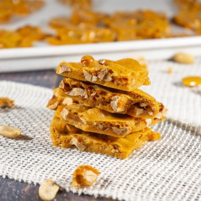 Four pieces of peanut brittle stacked on a burlap sheet on a wooden table with scattered peanuts and brittle around it