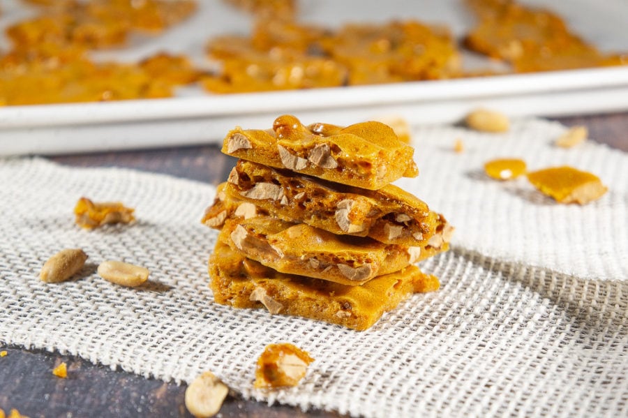 Four pieces of peanut brittle stacked on a burlap sheet on a wooden table with scattered peanuts and brittle around it