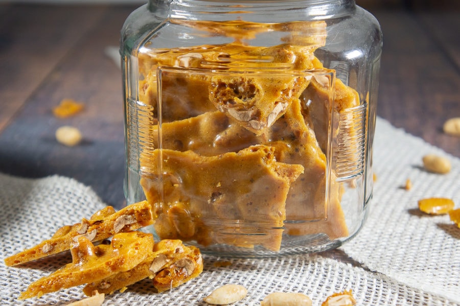 a jar of peanut brittle with a few pieces taken out on a wooden table.