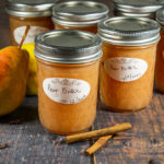 Six jars of pear butter on a wooden table with a whole pear and a few cinnamon sticks