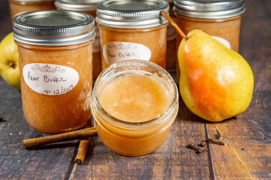 several unopened jars of pear butter with an open jar in front beside a fresh pear and cinnamon sticks
