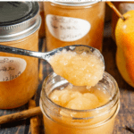 A small jar of pear butter open with a spoon dipping in and a few jars behind it and cinnamon sticks on the wooden table