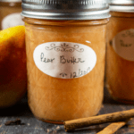 An 8 ounce jar of a pear butter recipe with two cinnamon sticks sitting beside it on a wooden table.