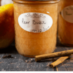 An 8 ounce jar of a pear butter recipe with two cinnamon sticks sitting beside it on a wooden table.