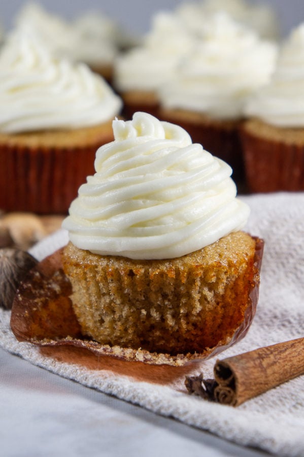 a spice cupcake with cream cheese frosting unwrapped on a grey towel beside a cinnamon stick in front of more cupcakes