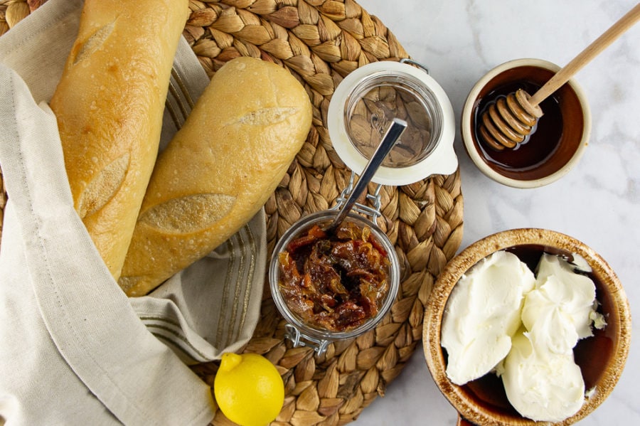 two baguettes with a bowl of creamy cheese, beside a jar of bacon jam, a bowl of honey, and a lemon