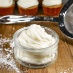 a bowl of cream cheese frosting in front of cupcakes
