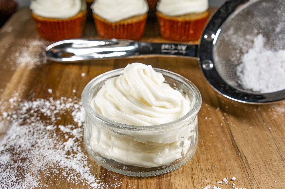 a bowl of cream cheese frosting in front of cupcakes