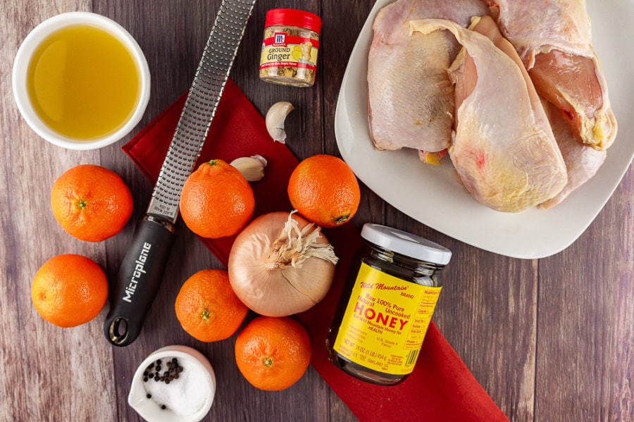 a plate of chicken beside a jar of honey, several mandarins, a cup of chicken broth, salt and pepper, onion, garlic, and ground ginger